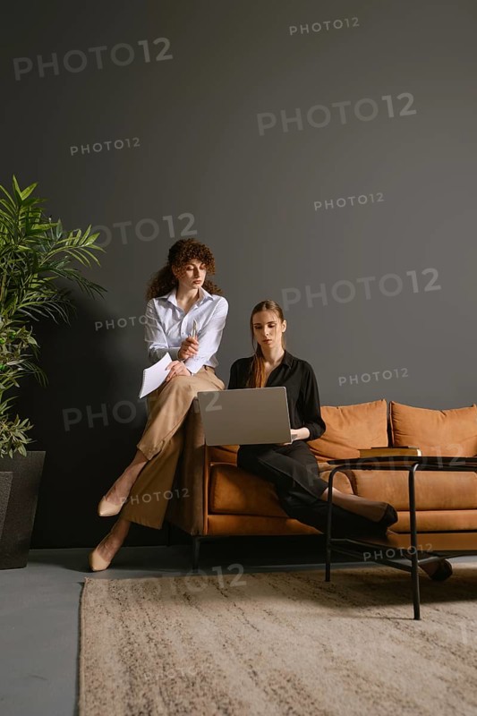 Two women focus on their work as they share ideas and discuss projects while seated in an office sofa. One woman stands beside the couch, and the other sits with a laptop, surrounded by greenery
