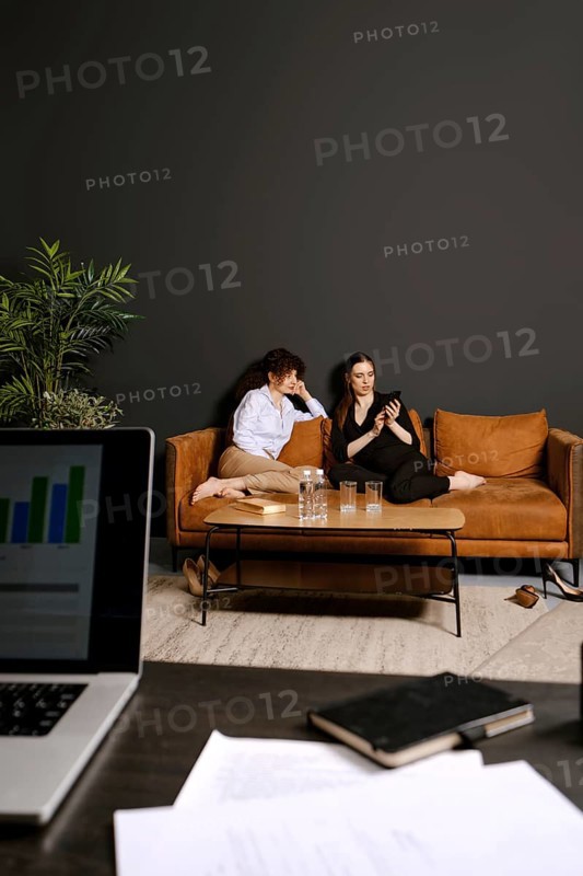 Two individuals are seated comfortably on a stylish couch, one looking at a phone while the other gazes thoughtfully. They are situated in a contemporary living room with a dark wall