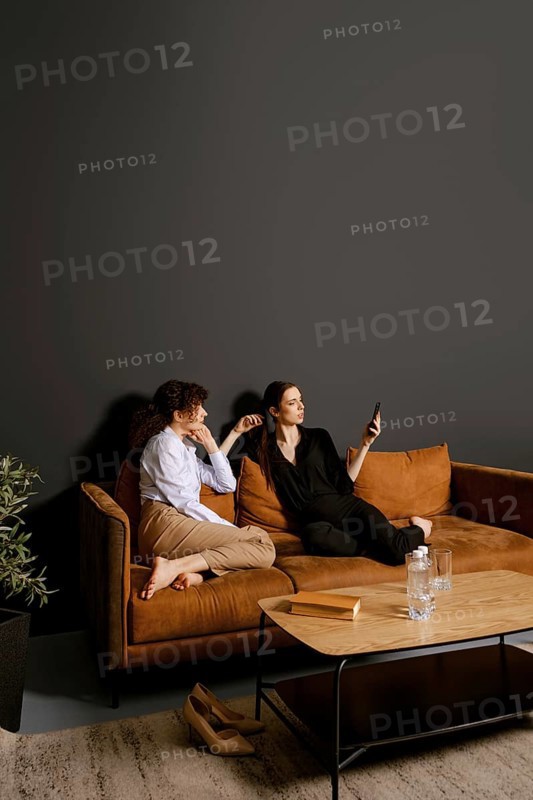 Two friends sit on a comfortable couch in a stylish living room. One is scrolling on a smartphone while the other watches with a relaxed expression, creating a warm and inviting atmosphere