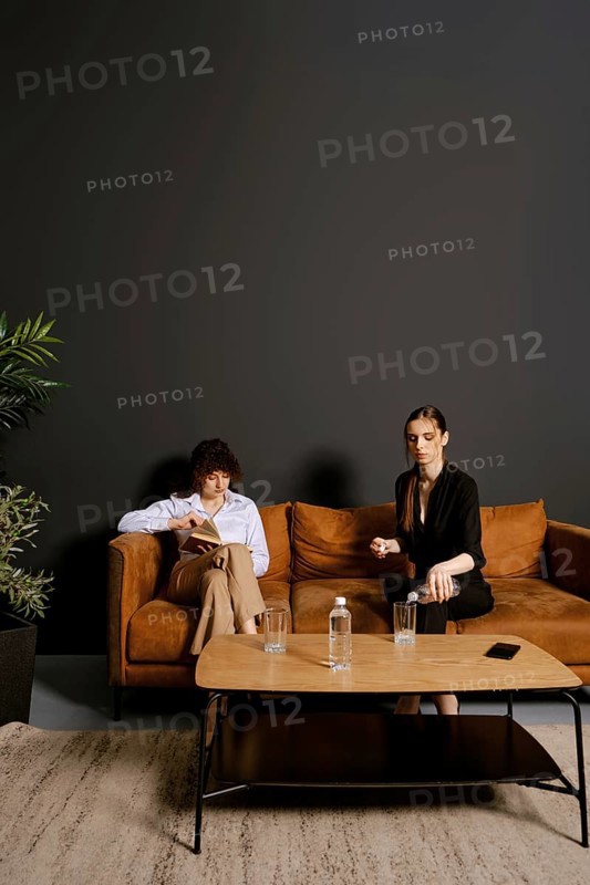 In a stylish indoor setting, two individuals sit on a brown couch. One person reads a book while the other holds a cup, engaged in conversation. A plant and water glasses complete the scene