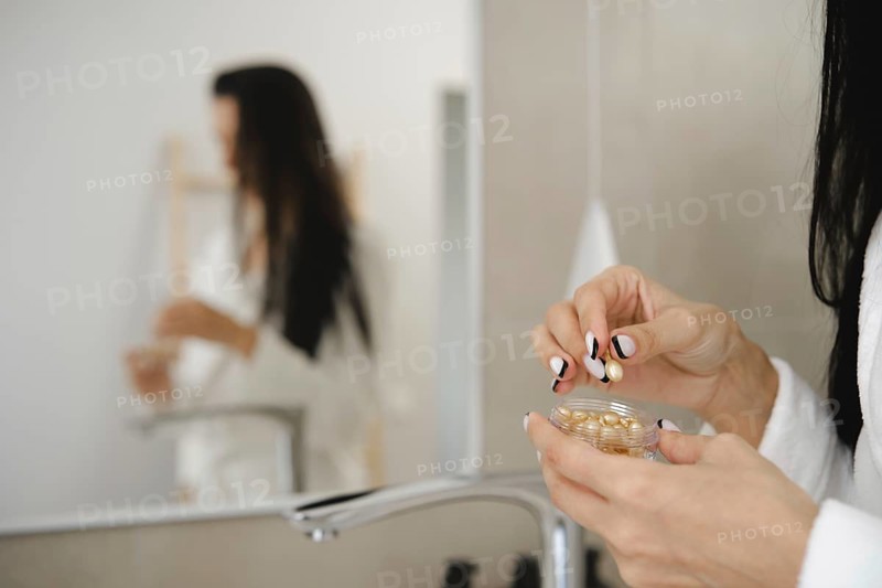 In a stylish bathroom, a woman in a robe is holding a small jar with skin vitamins and preparing to apply them