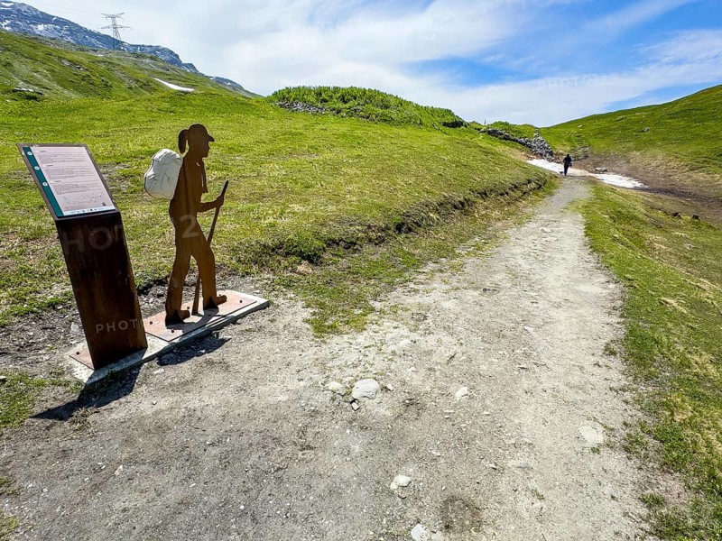 Information board on the left next to the Alpine Pass border pass road on early history and pass crossing in ancient times from the Alpine Pass border pass Little St. Bernard, next to it a stylised hiker with a walking stick and rucksack, on the right an old mule track parallel to the pass road, Aosta Valley region, Italy