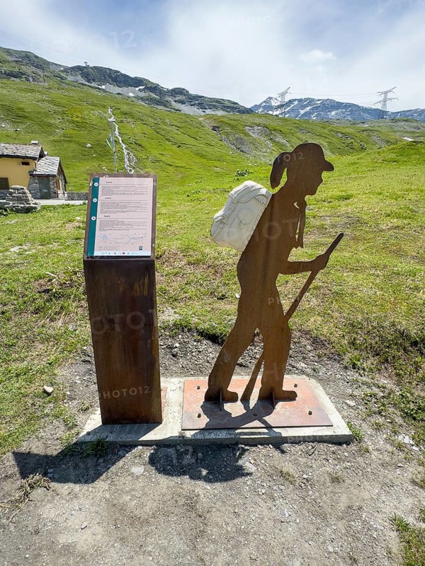 Information board on the left next to the Alpine Pass border pass road on early history and pass crossing in ancient times from the Alpine Pass border pass Little Saint Bernard, on the right stylised hiker with walking stick and rucksack, Aosta Valley region, Italy