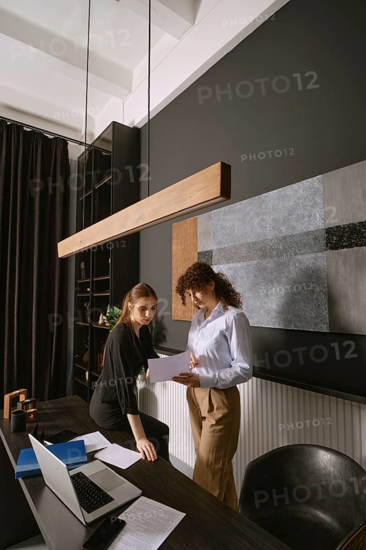 Two women engaged in conversation while reviewing documents in a stylish office. The setting features modern furniture and artistic wall decor. A laptop and papers are also visible on the desk