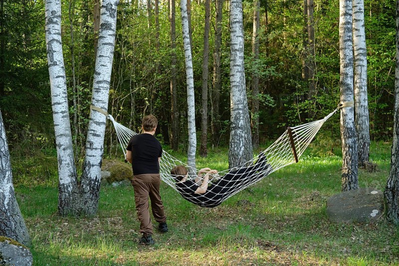 Two children enjoy their time outdoors, one relaxing in a hammock suspended between two trees while the other playfully holds onto the hammock