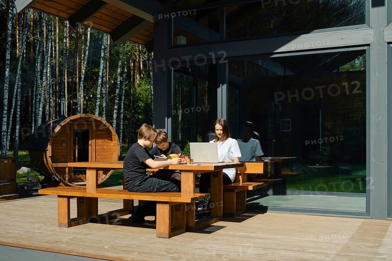 Two children focus on their tasks at a wooden table outside a modern cabin, while their mother guides them using a laptop. Surrounding trees offer a tranquil ambiance perfect for learning