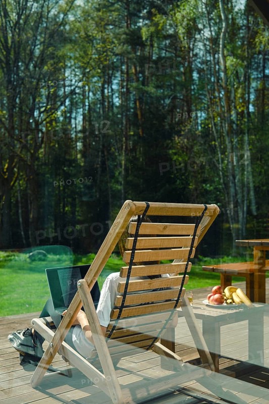 A person enjoys leisure time on a wooden lounge chair, surrounded by picturesque trees and a calm atmosphere. Refreshing fruits lie on a nearby table, enhancing the peaceful outdoor experience