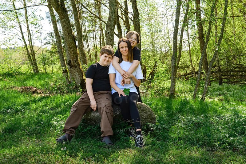 A single mother sits comfortably on a large rock with her two sons in a vibrant green forest clearing. They stopped to rest after a walk in the woods