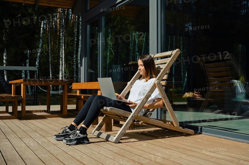 A woman sits comfortably in a wooden chair on a deck, focused on her laptop. Bright sunlight filters through trees, creating a serene atmosphere perfect for remote work or relaxation