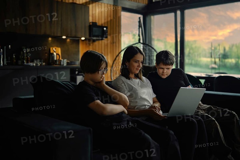 Young mother and her sons watch a series on a laptop sitting in the evening in a cozy living room of a wooden cabin