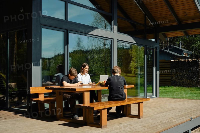 A family sits together at a wooden table on a sunny afternoon, engaged in various activities. One child uses a smartphone, while another focuses on a laptop, surrounded by nature