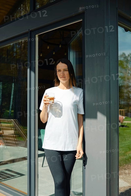 A young woman stands leisurely at the entrance of a stylish house, holding a drink in her hand. Sunlight shines through the glass doors, creating a warm atmosphere in a green backyard