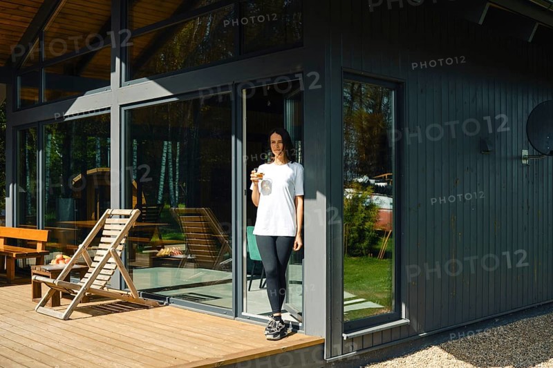 A woman stands on the deck of a stylish house, holding a drink while enjoying a sunny day. The home features large windows and a relaxing outdoor area with furniture, surrounded by greenery
