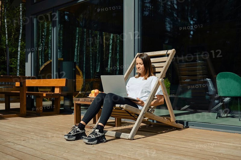 A woman is sitting in a wooden lounge chair, engaged with her laptop on a sunny deck. She enjoys a relaxed atmosphere surrounded by greenery, with fresh fruit nearby and large windows in view