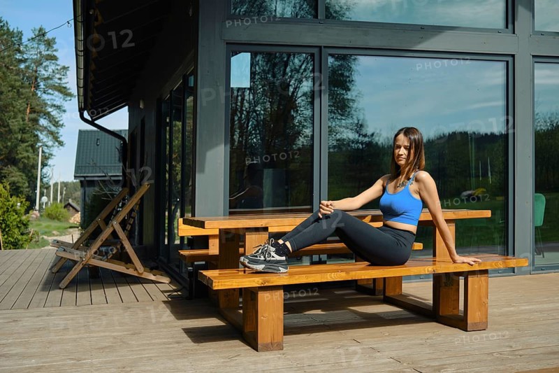 A woman is seated on a wooden bench, stretching her leg while practicing yoga in a serene outdoor setting