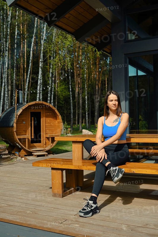 A woman dressed in casual sportswear sits comfortably on a wooden bench in front of a traditional sauna. Surrounded by tall trees and greenery, the day is bright and inviting, perfect for unwinding