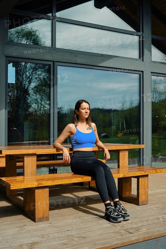 A woman sits on a wooden bench in front of a large glass cabin, basking in natural light. Surrounded by greenery, she wears a blue top and black leggings, appearing relaxed and peaceful