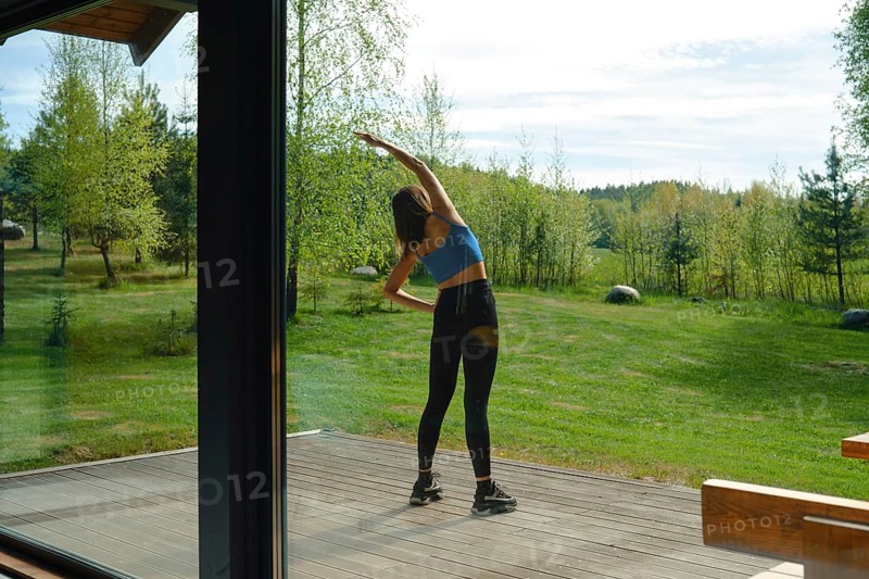 The view from inside the house at a woman is stretching her body on a wooden deck surrounded by lush green trees on a sunny day. The natural setting provides a peaceful backdrop for her fitness activity