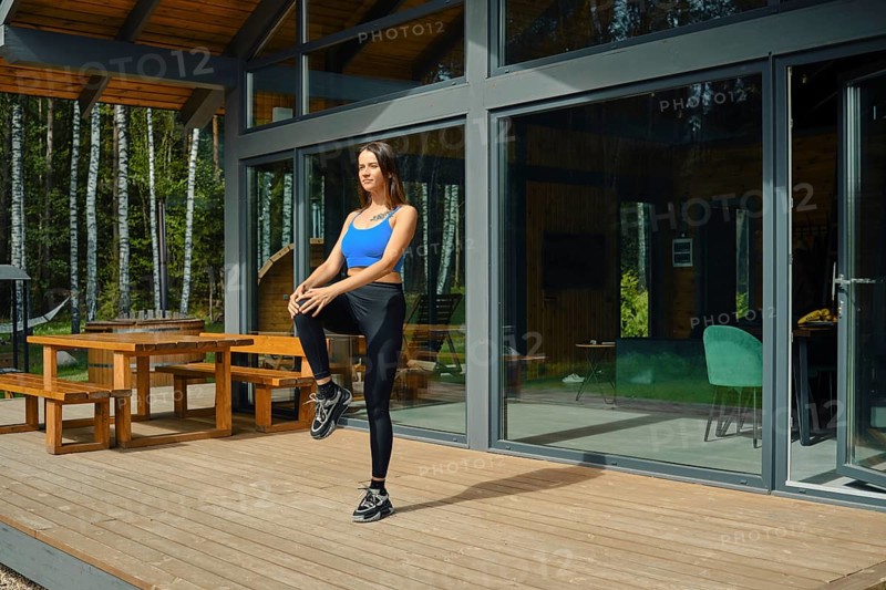 A woman wearing athletic attire is engaged in a warm-up exercise, raising one knee while standing on a wooden deck outside a contemporary cabin. Lush green trees provide a peaceful backdrop