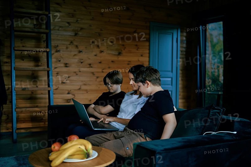 A family sits together on a comfortable couch in a wooden living room, focusing on a laptop. The mother explains the rules and teaches the children using interactive learning