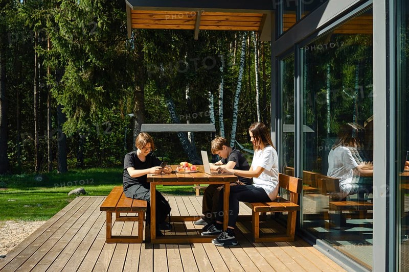 A single mother works remotely at a laptop sitting at a table on the terrace of a country house, and her sons are passionate about a game on a smartphone