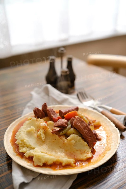 A hearty serving of creamy mashed potatoes topped with tender meat and vegetables on a rustic wooden table. Soft light illuminates the meal, enhancing the inviting atmosphere of the setting