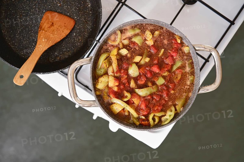 A pot filled with chopped vegetables and pork meat simmers on a stovetop. Fresh tomatoes, green peppers, and spices create a colourful and aromatic stew