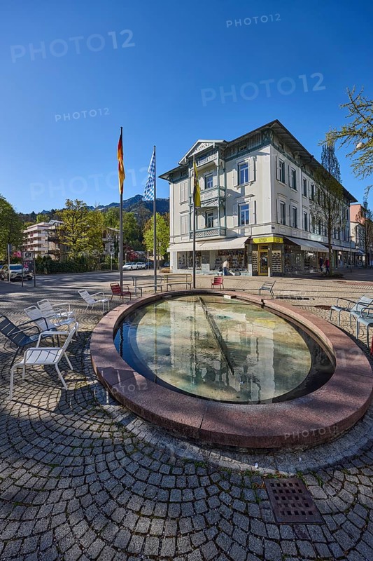 Wisbacherbrunnen, fountain, public chairs, flagpoles, flags, German flag, Bavarian flag, pedestrian zone, retail shops, buildings, trees, blue sky, cloudless, Wisbacherstrasse, Ludwigstrasse, Salzburger Strasse, Bad Reichenhall, Berchtesgadener Land district, Bavaria, Germany