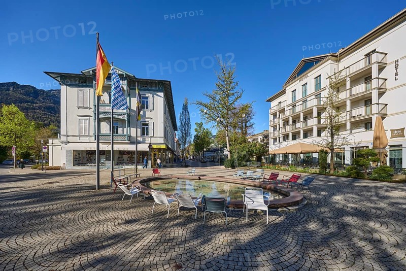 Wisbacherbrunnen, fountain, public chairs, flagpoles, flags, German flag, Bavarian flag, building, hotel, ibis Styles, trees, blue cloudless sky, Ludwigstrasse, Wisbacherstrasse, Salzburger Strasse, Bad Reichenhall, Berchtesgadener Land district, Bavaria, Germany