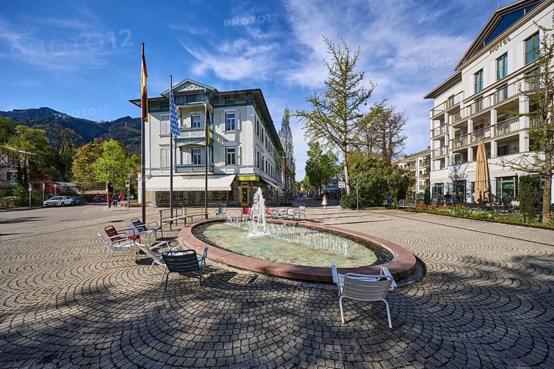 Wisbacherbrunnen, fountain, pedestrian zone, general architecture, cobblestones, flagpoles, flags, trees, blue sky, cirrostratus clouds, crossing of Salzburger Straße with Wisbacher Straße and Ludwigstraße, Bad Reichenhall, district Berchtesgadener Land, Bavaria, Germany