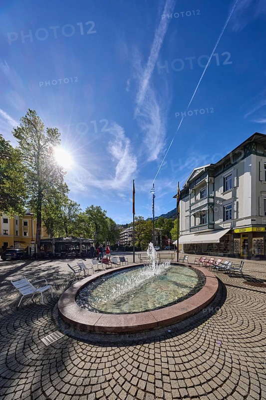 Wisbacherbrunnen, fountain, pedestrian zone, general architecture, cobblestones, public chairs, flagpoles, flags, trees, backlight of the sun, blue sky, cirrostratus clouds, intersection of Salzburger Straße with Wisbacher Straße and Ludwigstraße, Bad Reichenhall, district Berchtesgadener Land, Bavaria, Germany