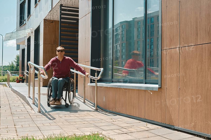 Individual in a wheelchair is utilizing a ramp to exit a modern building with large windows. The bright sun illuminates the exterior, creating a vibrant urban atmosphere