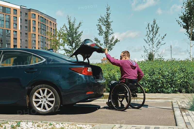 A man in a wheelchair is placing items into the trunk of a car in a bright urban area. The scene shows modern buildings and greenery, highlighting accessibility and independence on a clear day