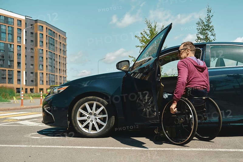 A man in a wheelchair is using his arms to propel himself into a parked vehicle with an open door. The backdrop features a contemporary apartment building under a clear blue sky