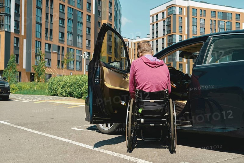 A man in a wheelchair is preparing to enter a car parked in a modern urban parking lot