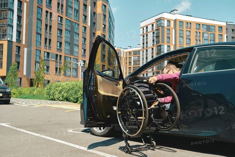 A person is maneuvering a wheelchair into a car parked in a contemporary urban area. A man unfolds a wheelchair to get into it from the car