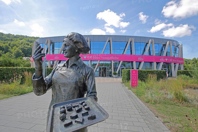 Sculpture confectioner with tray full of pralines for tasting, presentation, eating, enjoyment, pleasure, look, marvel, look at, enjoy, modern, round, building, glass window, pink banner with inscription, anniversary 130 years of nougat love, chocolate factory, sale, production and manufacture of sweets, chocolate, nougat, confectionery, confectioner, chocolate, food, Viba Nougat World, Schmalkalden, Franconia, Thuringia, Germany