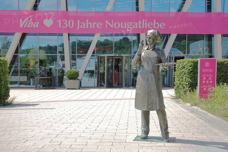 Sculpture confectioner with tray full of pralines for tasting, presentation, eating, enjoyment, pleasure, look, marvel, look at, enjoy, modern, round, building, glass window, pink banner with inscription, anniversary 130 years of nougat love, food, chocolate factory, sale, production and manufacture of sweets, chocolate, nougat, confectionery, confectioner, chocolate, entrance, Viba Nougat World, Schmalkalden, Franconia, Thuringia, Germany