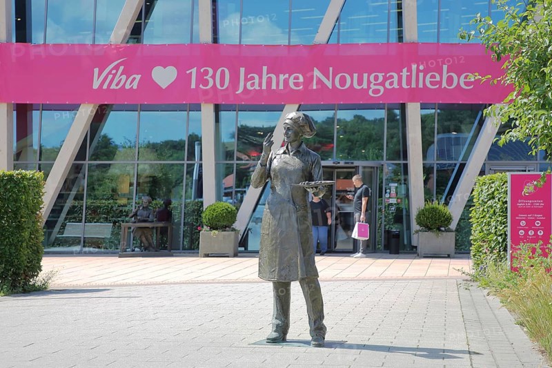 Sculpture confectioner with tray full of pralines to taste, presentation, eat, enjoyment, pleasure, look, marvel, look at, enjoy, modern, round, building, glass window, pink banner with inscription, anniversary 130 years of nougat love, chocolate factory, sale, production and manufacture of sweets, chocolate, nougat, confectionery, confectioner, chocolate, entrance, visitor, Viba Nougat World, Schmalkalden, Franconia, Thuringia, Germany