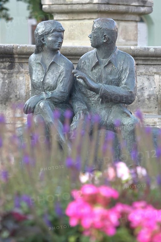Bronze sculpture at the market fountain, modern art, male figure, female figure, couple, sitting, conversation, entertainment, talking, listening, looking, joy, joyful, friends, relationship, communication, friendship, partnership, floral decoration, depth of field, blur, Schleusingen, Thuringia, Germany