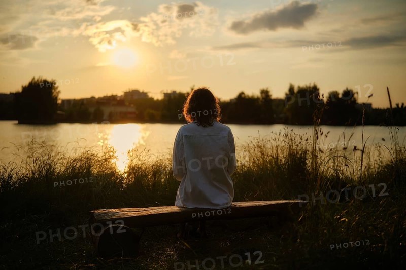 At the edge of a serene lake, a woman sits on a wooden bench surrounded by tall grass, soaking in the warm sun and vibrant colors of the sunset