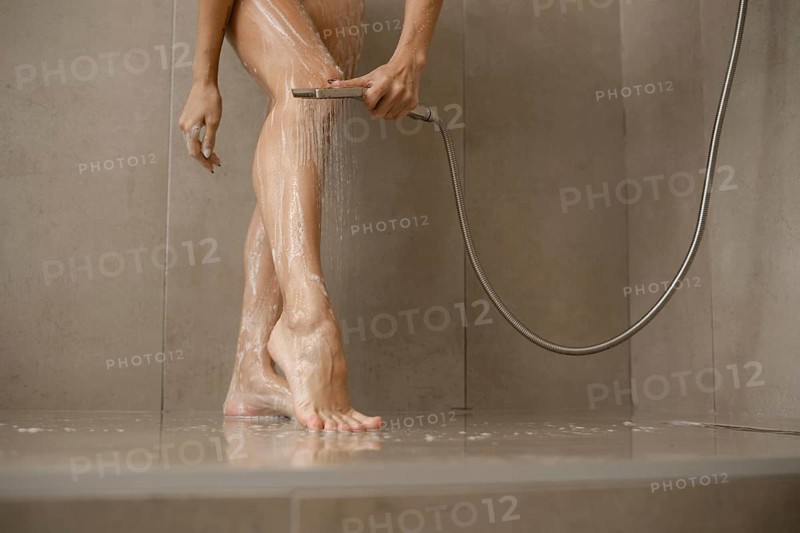 Water cascades over female legs in a sleek bathroom. The individual uses a handheld showerhead to clean and refresh while standing on a tiled floor