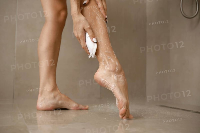 A woman is focused on washing her leg in a bathroom. The soft natural light enhances the clean setting, with water droplets glistening on her skin and the tiled floor