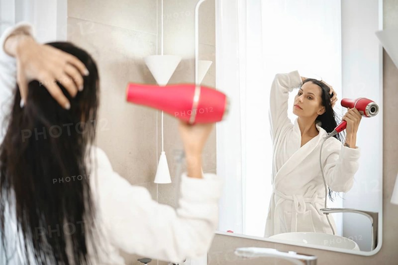 A woman is using a hair dryer to style her wet hair while standing in front of a large mirror. The bright bathroom features minimalistic design elements and soft natural light filtering through