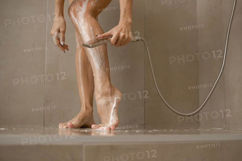 A woman stands in a sleek shower, washing soap off her legs while using a handheld showerhead. Water cascades around her feet, showcasing a fresh start to the day in a serene bathroom setting