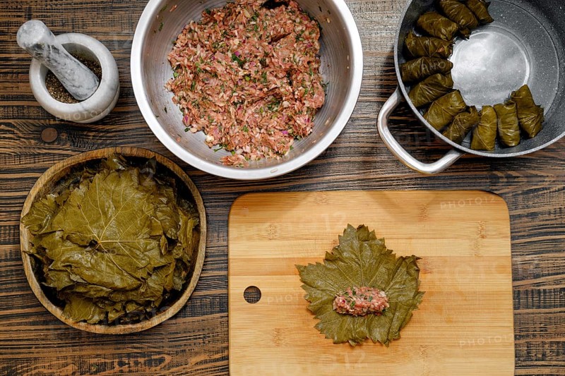 In a cozy kitchen, grape leaves are prepared for making dolma. A mixture of rice and minced meat is being formed into small portions on a wooden board, ready for wrapping in leaves