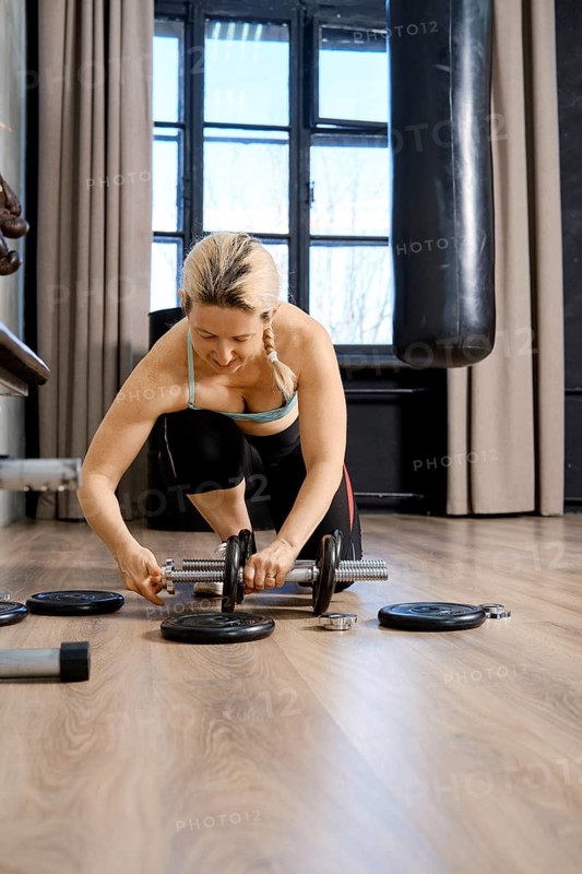 A focused woman is adjusting weights while kneeling on a gym floor. The space has large windows allowing natural light to illuminate the workout area, creating an inspiring atmosphere