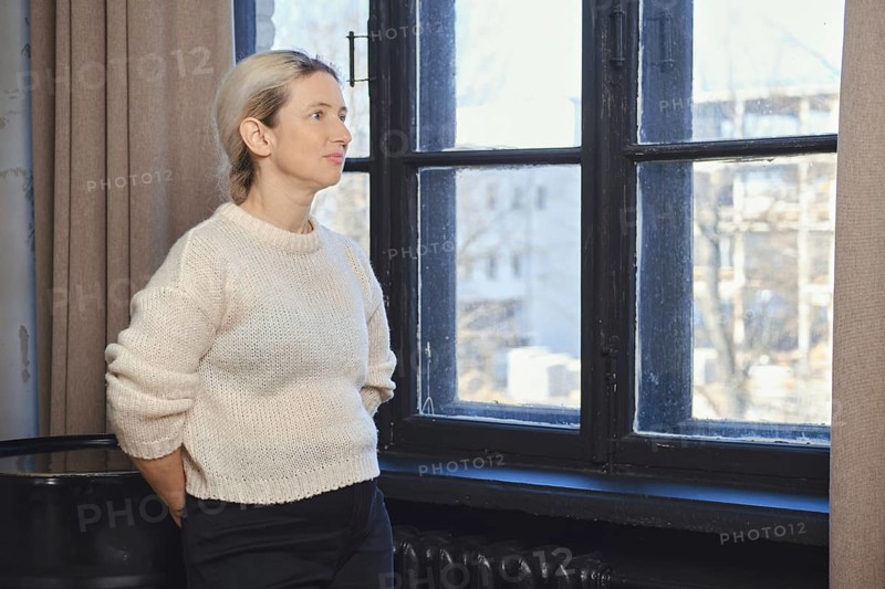 A woman stands next to a large window, lost in thought as she gazes outside. The sunlight filters into the room, highlighting her soft sweater and the serene atmosphere
