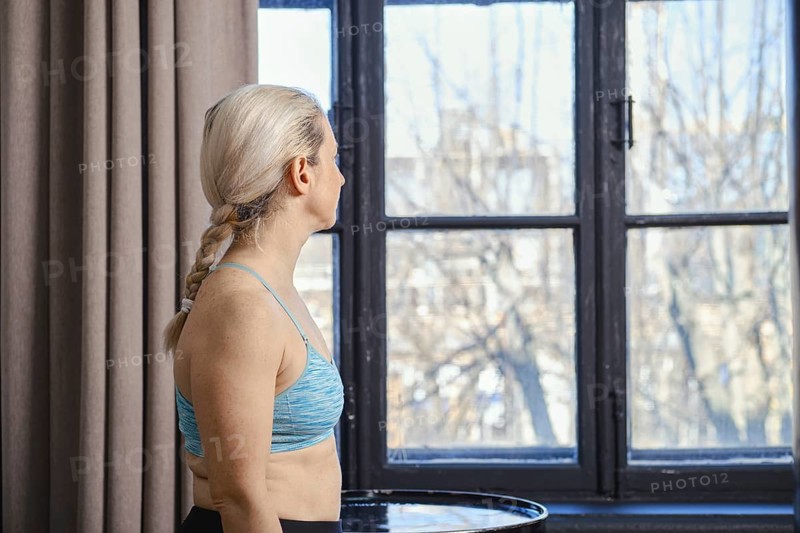 A woman with braided hair is standing near a large window, looking outside at the bright day. She wears a blue athletic top and appears thoughtful, surrounded by soft curtains and natural light