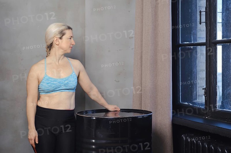 A woman wearing a blue sports bra and black leggings stands next to a black barrel, gazing thoughtfully out the window. The minimalist room features a neutral-toned wall and soft natural light